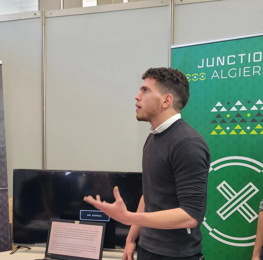 Man with short curly hair and a beard, wearing a dark sweater, mid-explanation with one hand raised while presenting at a tech event booth. A laptop showing project notes about fire detection sits in front of a blank TV screen, with a green 'Junction Algiers' banner in the background